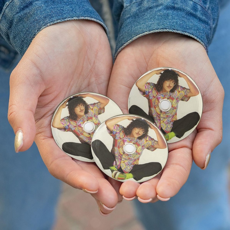 May include: Three round buttons featuring a person in a colorful shirt and black pants. The buttons are held in cupped hands against a denim background. Each button has a white center.
