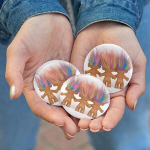 May include: Three round white buttons featuring a cartoon troll design with brown bodies and colorful rainbow hair. The buttons are held in open hands against a denim background. The buttons are approximately 1.5 inches in diameter.