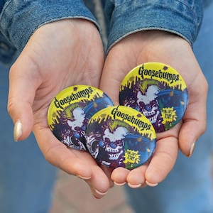 May include: Three yellow Goosebumps buttons with a spooky illustration of a skull-like character. The buttons have the text "Goosebumps" in a green and blue font. The buttons are held in open hands.