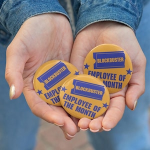 May include: Three round, yellow Blockbuster "Employee of the Month" pinback buttons. Each button features the Blockbuster logo in blue and the text "Employee of the Month" in blue. The buttons are held in open hands.
