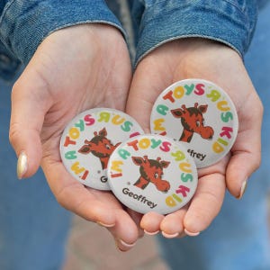 May include: Three round white buttons with the text "TOYS 'R' US KID" and a cartoon giraffe. The buttons are held in open hands. One button has the name "Geoffrey" and another has "'frey". The buttons have red, yellow, and green accents.