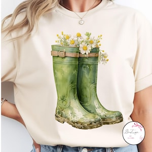 Puede incluir: Camiseta color crema con una ilustración en acuarela de botas de agua verdes llenas de flores blancas y amarillas. Las botas tienen correas y hebillas marrones. El diseño evoca un tema de primavera o jardín.