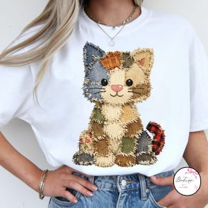 Op de afbeelding: Wit T-shirt met een patchwork kattenontwerp. De kat is gemaakt van verschillende stoffen patches in tinten blauw, bruin, groen en rood. De kat heeft een roze neus en een lachend gezicht. Het shirt is een casual stijl.