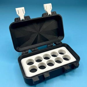 Könnte beinhalten: Offene, rechteckige schwarze Box mit einem weißen Inneneinsatz mit 10 kreisförmigen Aussparungen. Die Box hat zwei weiße Verschlüsse und befindet sich vor einem blauen Hintergrund. Die Box ist für kleine Gegenstände konzipiert.