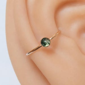 May include: A delicate gold-coloured ring featuring a small, round, green gemstone. The ring is thin and simple, with the gemstone set in a bezel setting. The ring is shown on a person's ear.