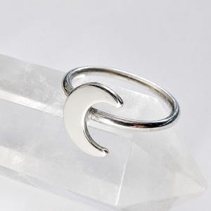 May include: A polished silver ring featuring a crescent moon design. The ring is resting on a clear, faceted quartz crystal point. The metal has a smooth, reflective surface, and the crescent moon shape is centered on the thin, circular band.