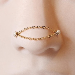 May include: A gold-toned nose chain featuring two delicate link chains that connect at each end to a small, round, white pearl-like bead. The jewelry piece is designed to drape across the bridge of the nose, creating a horizontal line.