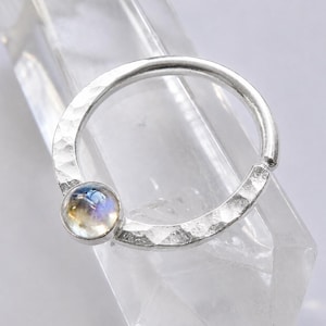Puede incluir: Un anillo circular de plata con una banda martillada y texturizada, que presenta un cabujón de piedra lunar redondo engastado en bisel. La piedra muestra una calidad translúcida e iridiscente con tonos azules y amarillos sobre la plata.