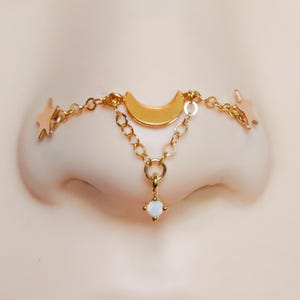 Puede incluir: Joya de nariz dorada con una luna creciente, estrellas y una piedra colgante con aspecto de ópalo. La delicada cadena conecta la luna y las estrellas, creando un accesorio de temática celestial. La joya está diseñada para ser usada en la nariz.
