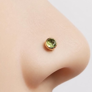 May include: A small, round nose stud featuring a bezel-set, faceted light green gemstone encased in a polished gold-toned metal setting. The circular stone is centered within the metal rim, creating a simple, low-profile design for facial jewelry.