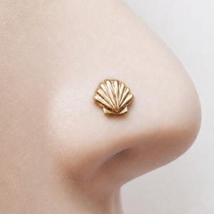 May include: A small, gold-toned nose stud featuring a detailed, fan-shaped seashell design. The jewelry piece has a polished metallic finish with embossed vertical ridges that mimic the natural texture of a scallop shell, centered on the nostril.