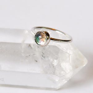 Peut inclure: Une bague en argent dotée d'une petite pierre circulaire sertie clos, avec une surface lisse et irisée présentant des éclats de couleurs vert, bleu et rose. L'anneau est fin et poli, arborant une finition métallique simple qui reflète la lumière.