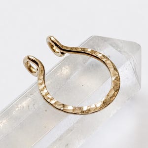 May include: A gold-toned, C-shaped nose cuff featuring a textured, hammered surface finish. The jewelry piece has two small, rounded loops at each end of the open band, creating a distinct, reflective appearance against the light.