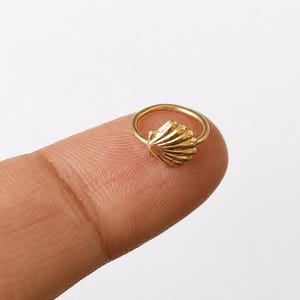 May include: A small gold hoop ring featuring a textured seashell charm. The circular band is thin and metallic, with a detailed, ridged fan-shaped shell element attached to the top, showcasing a polished finish and a distinct coastal aesthetic.