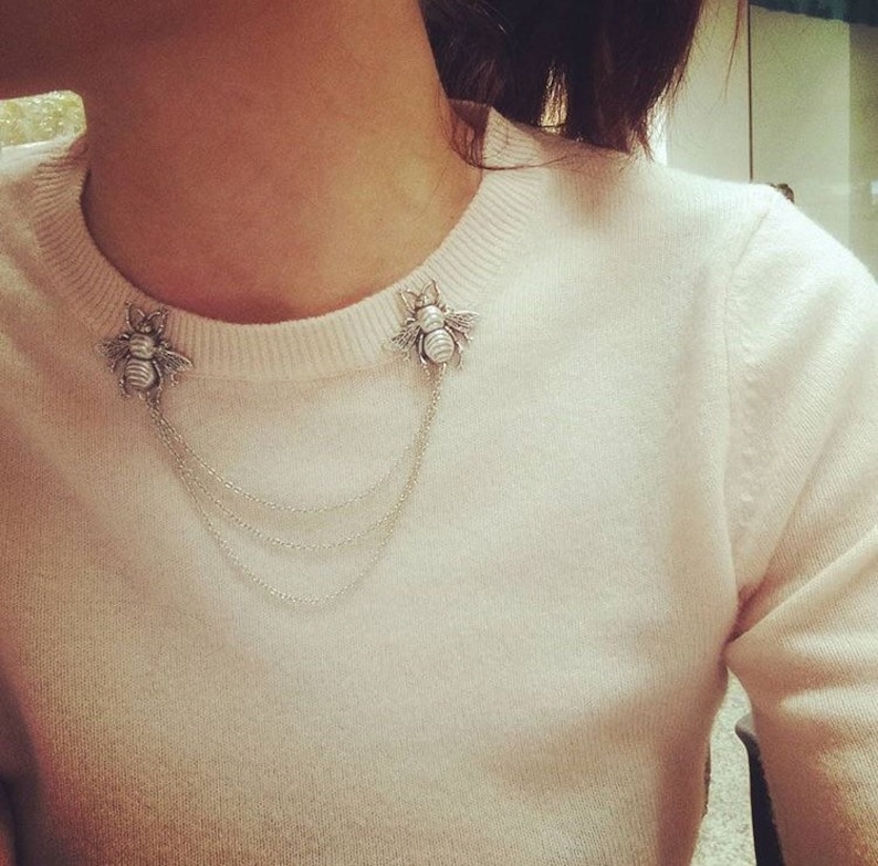 May include: A white sweater with a silver chain necklace featuring two bee-shaped pendants.