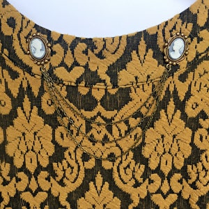 Op de afbeelding: Een paar gouden broche met cameo portretten, verbonden door een ketting, op een gouden en zwarte gepatroneerde stof.