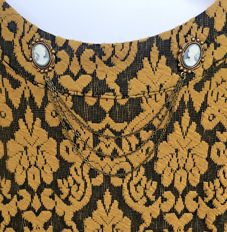 Op de afbeelding: Een paar gouden broche met cameo portretten, verbonden door een ketting, op een gouden en zwarte gepatroneerde stof.