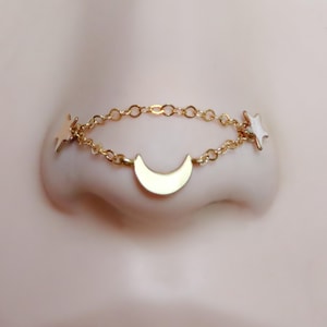 May include: A gold-colored nose chain featuring a crescent moon and two stars. The chain is delicate and connects to the nose piercing. The stars have white enamel detailing. The jewelry is designed to be worn on the nose.