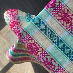 Puede incluir: Primer plano de un textil tejido colorido con un patr&oacute;n floral y geom&eacute;trico. La tela presenta rayas en tonos rosa, verde y turquesa, con motivos de rosas bordadas. El textil est&aacute; doblado, revelando su intrincado dise&ntilde;o.