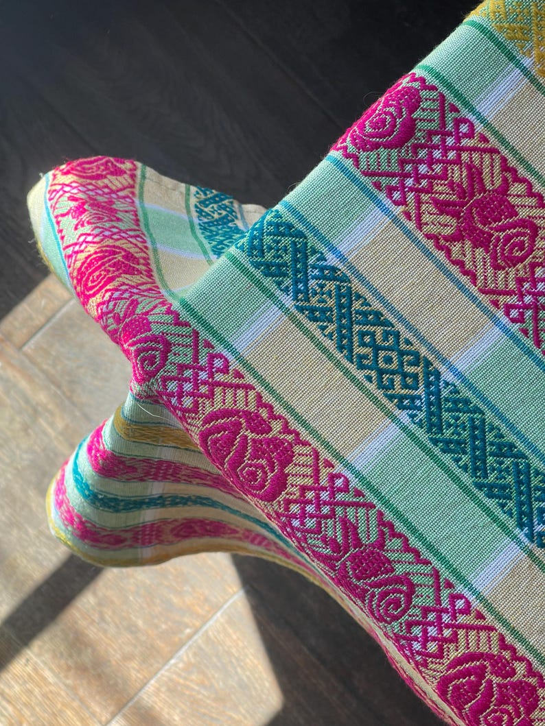 Puede incluir: Primer plano de un textil tejido colorido con un patr&oacute;n floral y geom&eacute;trico. La tela presenta rayas en tonos rosa, verde y turquesa, con motivos de rosas bordadas. El textil est&aacute; doblado, revelando su intrincado dise&ntilde;o.