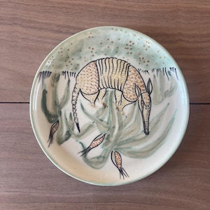 May include: A round ceramic plate featuring a hand-painted illustration of an armadillo in shades of tan and brown. The background is a pale green wash with dark green foliage accents and small gold-colored dots scattered across the upper portion.