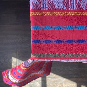 Puede incluir: Un textil rojo vibrante con intrincados patrones tejidos. El dise&ntilde;o presenta formas geom&eacute;tricas repetidas y figuras de animales en blanco y morado, con bandas azules, amarillas y verdes. La tela est&aacute; doblada, revelando un borde a rayas.