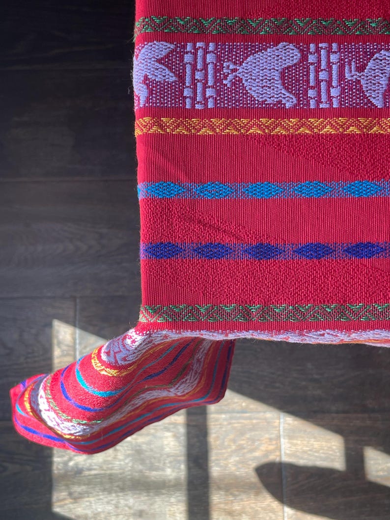 Puede incluir: Un textil rojo vibrante con intrincados patrones tejidos. El dise&ntilde;o presenta formas geom&eacute;tricas repetidas y figuras de animales en blanco y morado, con bandas azules, amarillas y verdes. La tela est&aacute; doblada, revelando un borde a rayas.