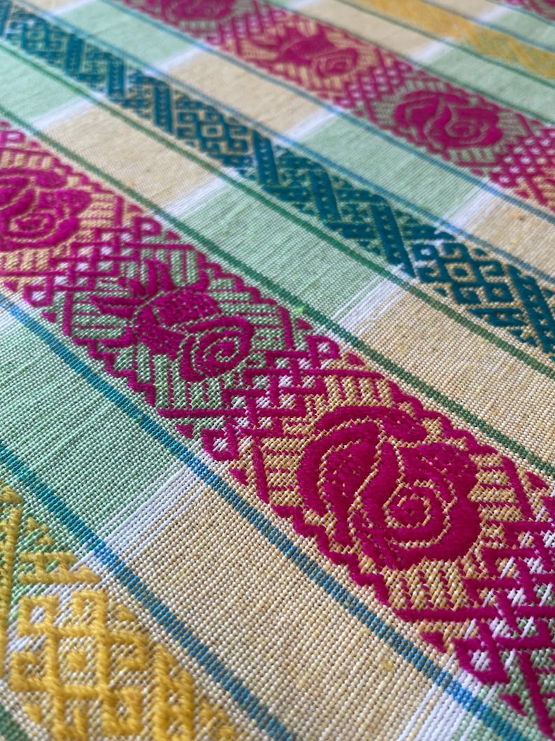 Puede incluir: Primer plano de un textil tejido con un patr&oacute;n repetitivo. La tela presenta rayas alternas amarillas, verdes y turquesas. Atrevidos dise&ntilde;os florales y geom&eacute;tricos fucsia est&aacute;n tejidos en la tela, creando una superficie texturizada.