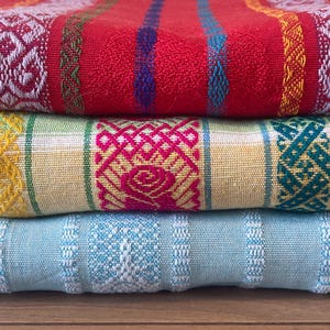 Puede incluir: Tres textiles plegados apilados. El textil superior es rojo con detalles blancos y coloridos. El textil del medio es amarillo con detalles rosas y verdes. El textil inferior es azul claro con detalles blancos.