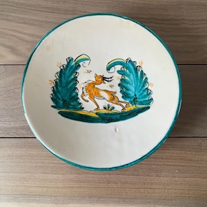 Peut inclure: Une assiette en céramique blanche avec un bord turquoise et un motif peint à la main. Le motif représente un lièvre brun courant entre deux plantes stylisées turquoise et jaunes. L'assiette est ronde et repose sur une surface en bois.