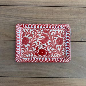 May include: A rectangular ceramic tray featuring a hand-painted red floral and bird motif on a white background. The border is decorated with a repeating red scalloped pattern, and the center displays a bird surrounded by stylized leaves and flowers.