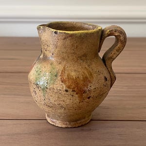 Puede incluir: Un pequeño jarro de cerámica de estilo antiguo con un cuerpo redondeado y un asa curvada. El jarro tiene una paleta de colores moteados y terrosos de marrones, beiges y toques de verde y amarillo, lo que sugiere una estética rústica o vintage.