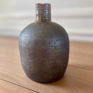 Puede incluir: Un jarrón de cerámica gris oscuro hecho a mano, con un cuerpo redondeado y un cuello cilíndrico texturizado. El jarrón tiene un acabado mate y una estética rústica, adecuado para la decoración del hogar.