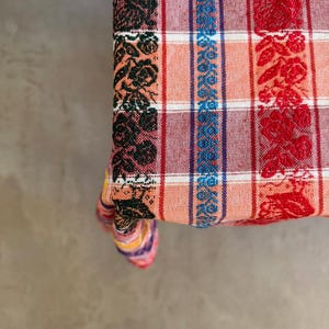 May include: A woven fabric textile featuring a plaid pattern in shades of orange, white, and dark red. The fabric is decorated with vertical bands of floral motifs in black, blue, and red, creating a textured appearance across the surface.