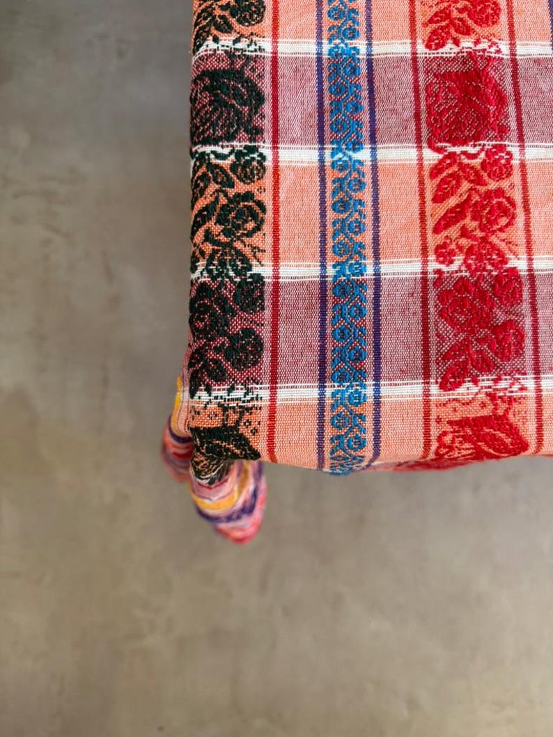 May include: A woven fabric textile featuring a plaid pattern in shades of orange, white, and dark red. The fabric is decorated with vertical bands of floral motifs in black, blue, and red, creating a textured appearance across the surface.