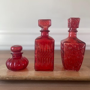 May include: Three red glass decanters of varying sizes. The decanters have decorative stoppers and textured surfaces. The decanters are arranged on a wooden tray. The decanters are likely for storing and serving beverages.