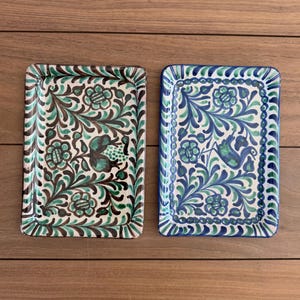 May include: Two rectangular ceramic trays with hand-painted floral and leaf patterns. The left tray features brown and green pigments on a cream base, while the right tray displays blue and green designs on a cream base, both with scalloped borders.
