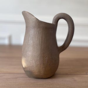 Puede incluir: Una pequeña jarra de cerámica marrón con un cuerpo redondeado y un asa curvada. La jarra tiene un pico para verter fácilmente y una superficie texturizada. El color neutro y el diseño simple la convierten en una pieza versátil para cualquier cocina o comedor.