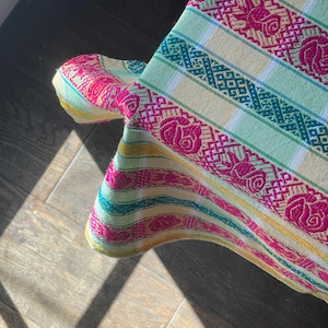 Puede incluir: Primer plano de un textil tejido colorido con un patr&oacute;n floral. La tela presenta rayas en tonos verdes, amarillos y rosas. El dise&ntilde;o incluye motivos de rosas repetidos y patrones geom&eacute;tricos, creando un aspecto vibrante y texturizado.