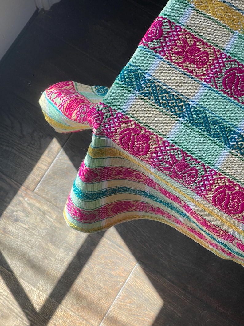 Puede incluir: Primer plano de un textil tejido colorido con un patr&oacute;n floral. La tela presenta rayas en tonos verdes, amarillos y rosas. El dise&ntilde;o incluye motivos de rosas repetidos y patrones geom&eacute;tricos, creando un aspecto vibrante y texturizado.