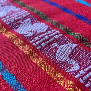 Puede incluir: Primer plano de un textil rojo tejido con rayas bordadas blancas, amarillas, verdes y azules. El textil presenta figuras blancas repetidas con forma de p&aacute;jaro y patrones geom&eacute;tricos, mostrando un dise&ntilde;o vibrante y detallado.