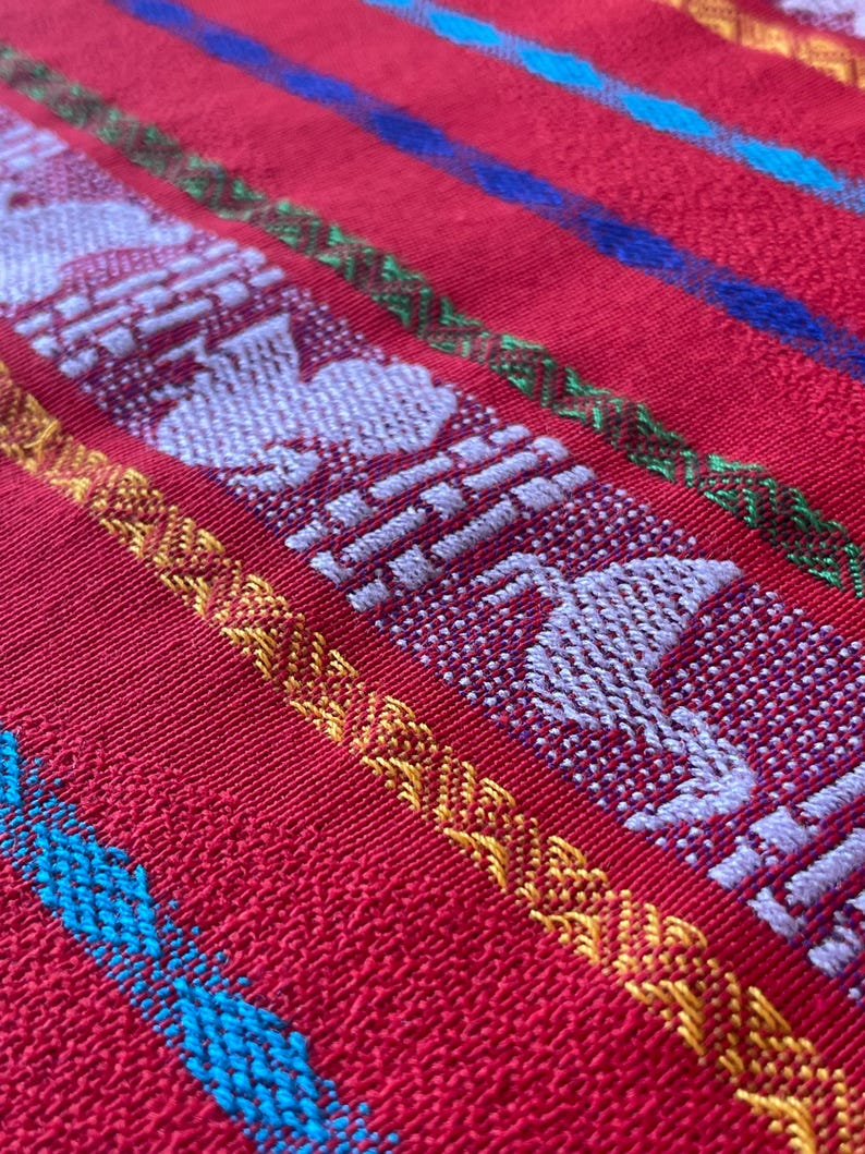 Puede incluir: Primer plano de un textil rojo tejido con rayas bordadas blancas, amarillas, verdes y azules. El textil presenta figuras blancas repetidas con forma de p&aacute;jaro y patrones geom&eacute;tricos, mostrando un dise&ntilde;o vibrante y detallado.