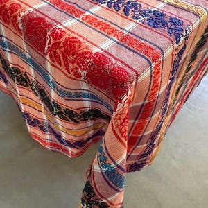 May include: A woven tablecloth featuring a plaid pattern in shades of peach and white, accented by vertical bands of raised floral embroidery in vibrant colors including red, blue, black, and yellow, draped over a square corner surface.
