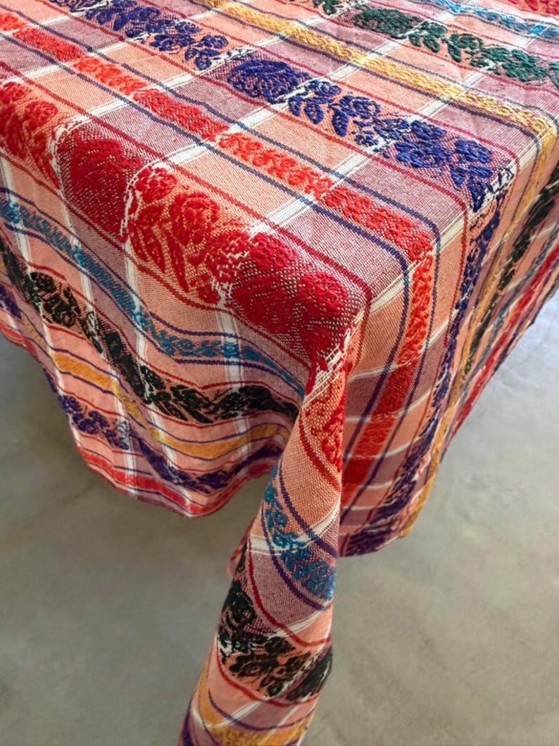 May include: A woven tablecloth featuring a plaid pattern in shades of peach and white, accented by vertical bands of raised floral embroidery in vibrant colors including red, blue, black, and yellow, draped over a square corner surface.