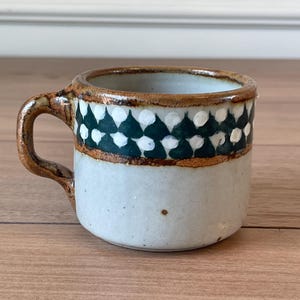 Könnte beinhalten: Eine kleine Keramiktasse mit Henkel. Die Tasse ist hellblau mit einem Band aus dunkelgrünen und weißen rautenförmigen Mustern am Rand. Der Rand und der Henkel sind braun.
