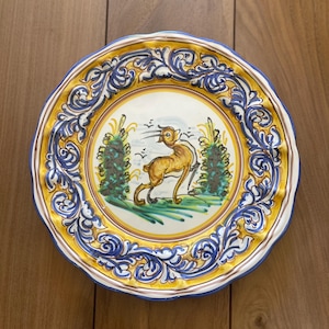 May include: A decorative ceramic plate with a hand-painted design. The plate features a central image of a deer in a landscape, surrounded by a yellow border and ornate blue and white scrollwork. The rim has a scalloped edge.