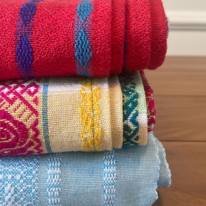 Puede incluir: Una pila de tres textiles tejidos doblados. El textil superior es rojo con detalles azules y turquesas. El textil del medio es amarillo con dise&ntilde;os rosas y verdes. El textil inferior es azul claro con detalles blancos. Los textiles est&aacute;n apilados sobre una superficie de madera.