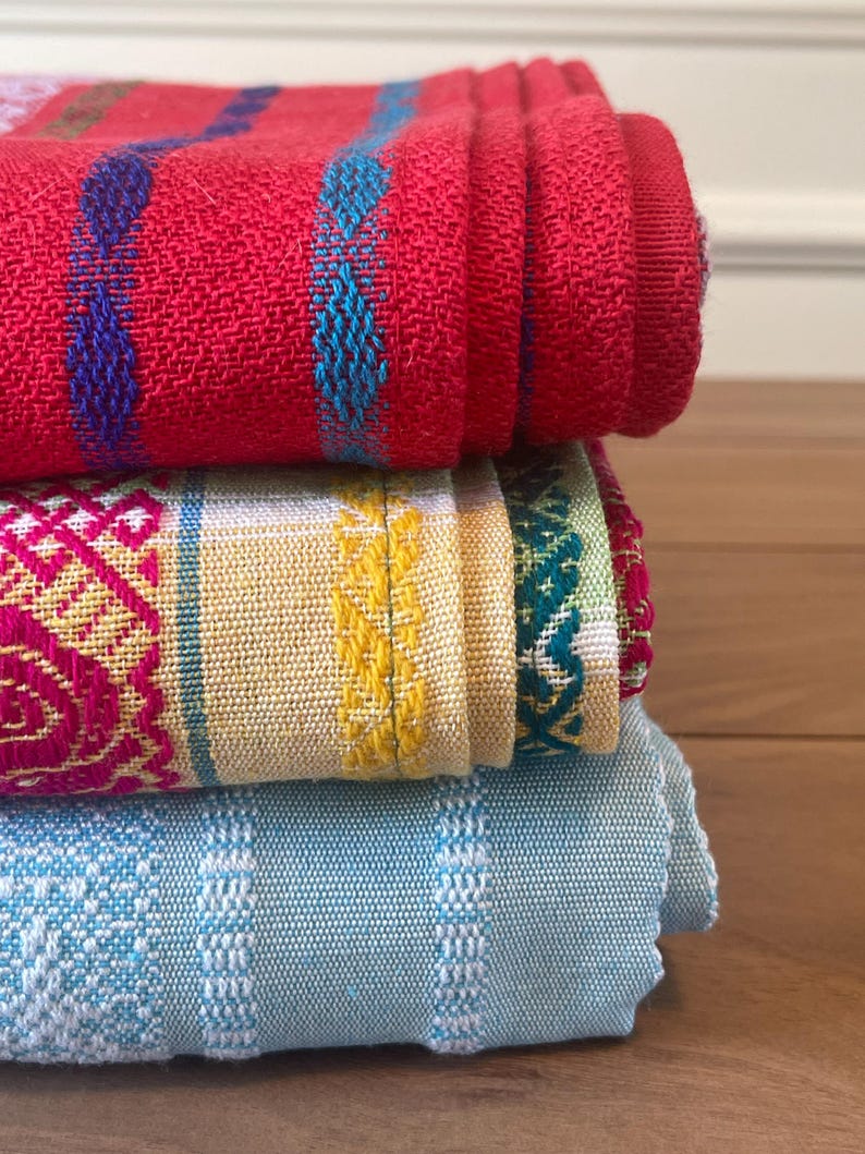 Puede incluir: Una pila de tres textiles tejidos doblados. El textil superior es rojo con detalles azules y turquesas. El textil del medio es amarillo con dise&ntilde;os rosas y verdes. El textil inferior es azul claro con detalles blancos. Los textiles est&aacute;n apilados sobre una superficie de madera.