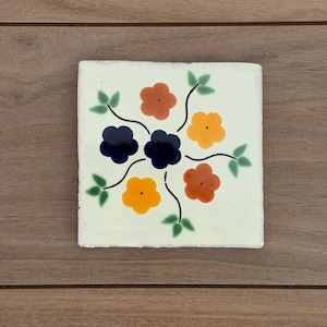 May include: A square ceramic tile with a cream-colored background featuring a central floral design. The pattern consists of six hand-painted flowers in shades of dark blue, orange, and yellow, connected by thin black stems with small green leaves.