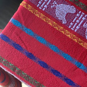 Puede incluir: Textil rojo tejido con rayas coloridas y patrones detallados. El dise&ntilde;o incluye figuras de animales blancos y formas geom&eacute;tricas, con acentos en azul, amarillo y verde. La tela tiene una apariencia texturizada.